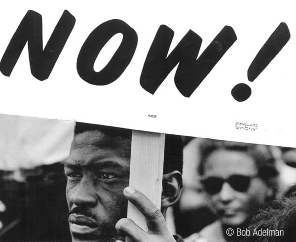 Black & white photograph of African-American man carrying a sign reading “Now”