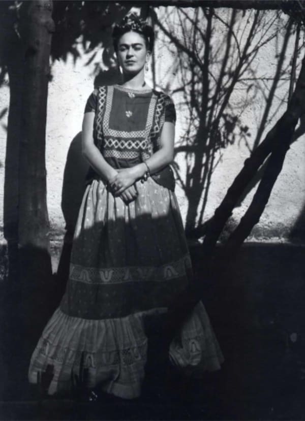 Black and white photograph of artist Frida Kahlo in front of an exterior wall of her home and studio, the Blue House, in between shadows cast by trees