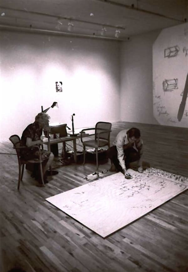 Black and White photograph of a man on the floor placing marks on a large map with a man sitting behind him looking at what he is doing.