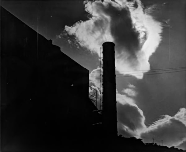Black and white photograph of the silhouette of a smoke stack against a cloudy sky