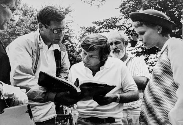 Black and white photograph of Roman Polanski reviewing the script with Mia Farrow and William A. Fraker in the background, outside in New York City, on the set of “Rosemary's Baby”