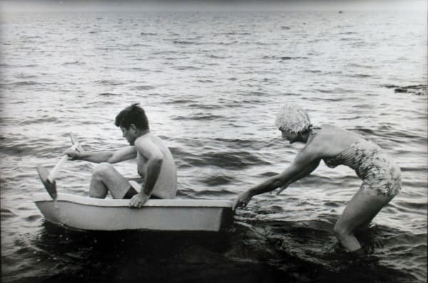 Black and white photograph of Jackie pushing John F Kennedy in a small boat in the water at a Hyannis Port beach