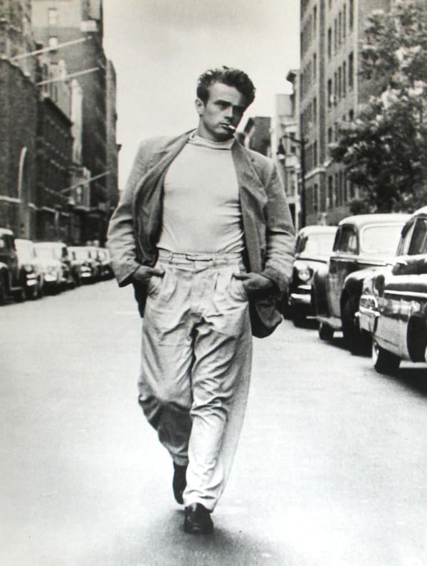 Black and white photograph of James Dean walking down a New York City street wearing a jacket, white shirt, and light pants, with a cigarette in his mouth