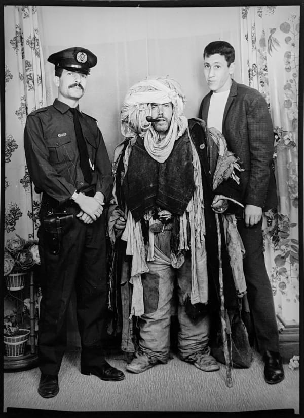 1960s black & white photograph of a policeman, a man wearing tribal garb and one man in western garb, standing in a photo studio
