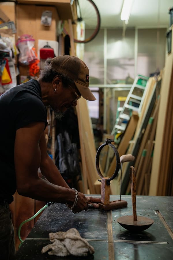 An artist, Alex Locadia, stands at a table working on a wood and metal sculpture amidst an industrial loft setting filled with materials and tools