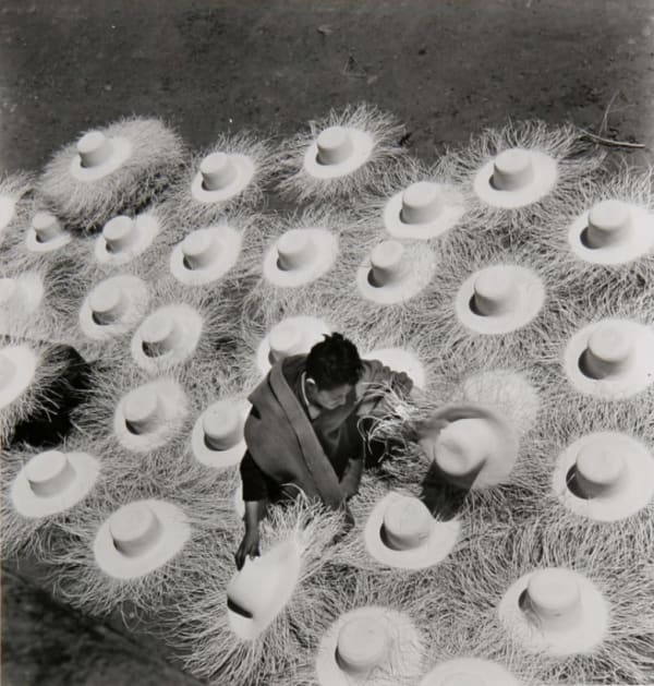Black and white photograph of a man walking amidst a multitude of palm hats arranged on the ground, taken from a high vantage point