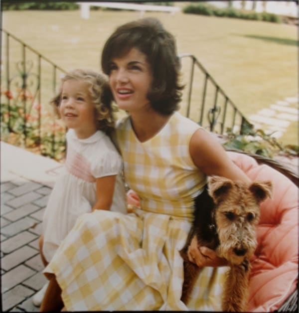 Color photograph of Jackie and a young Caroline Kennedy sitting on a patio with a terrier dog on a summer day