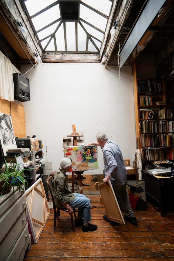 An artist Ken Jacobs standing and artist Flo Jacobs sitting at an easel with an unfinished painting in a loft setting filled with books and illuminated by a skylight