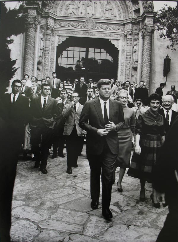 Black and white photograph of John F Kennedy walking toward the camera in front of a crowd of people and an ornate architectural façade in Paris