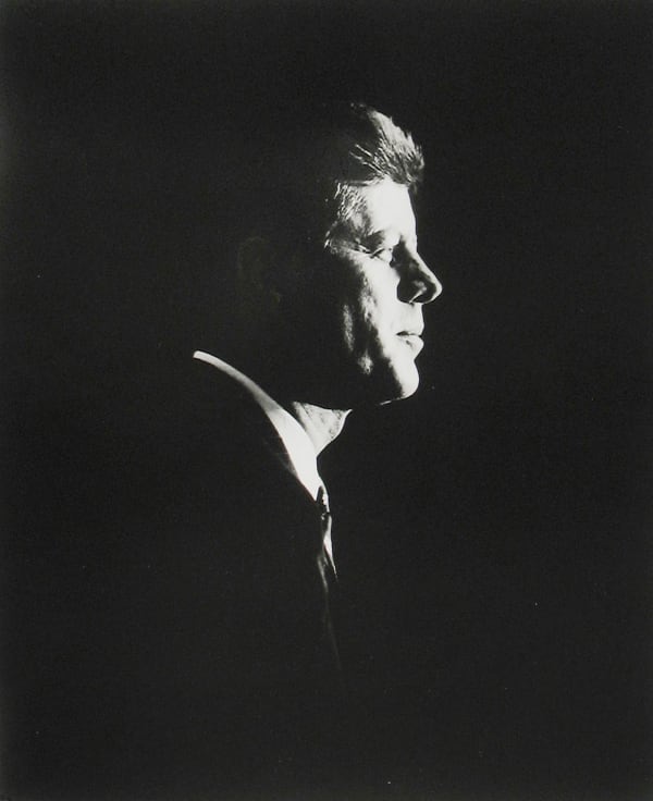 Black and white, high contrast photograph of John F Kennedy in profile against a pitch dark background