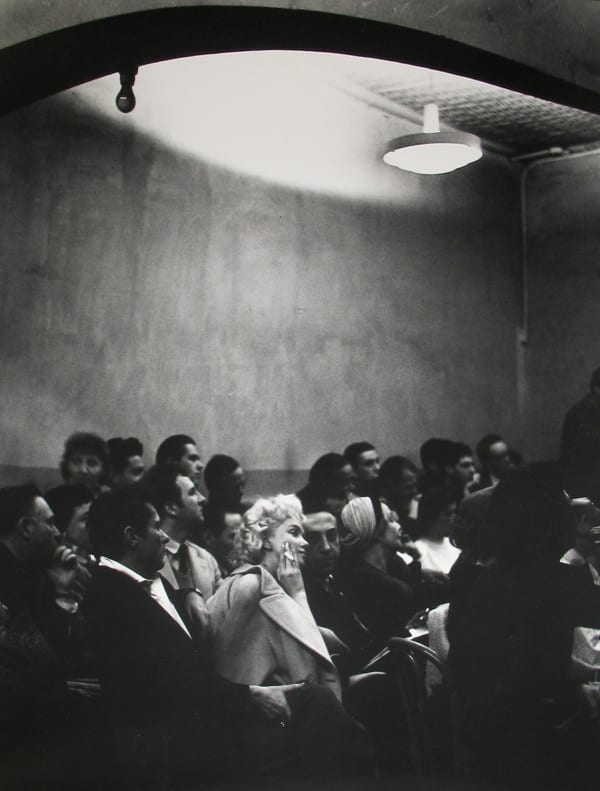 Black and white photograph of Marilyn Monroe holding a cigarette in a light-colored coat sitting amongst a crowd of other figures dressed in dark colors within a studio setting