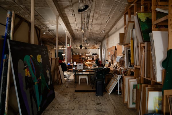 An artist, Carmen Cicero, sits at a desk working on an artwork in an industrial loft setting filled with paintings