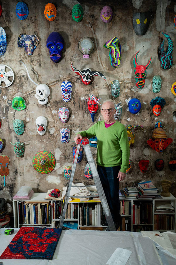 An artist, Jeff Way, poses on a ladder in front of a wall of colorful masks in a loft setting