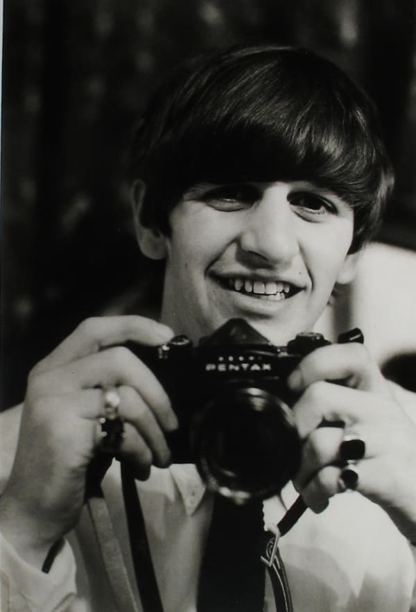 Black and white photograph of George Harrison holding a camera and smiling