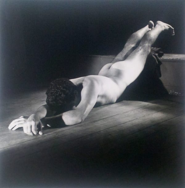 Black and white photograph of a naked man lying face down on a wooden floor under a spotlight, with his legs elevated on a step, and wrists crossed over one another