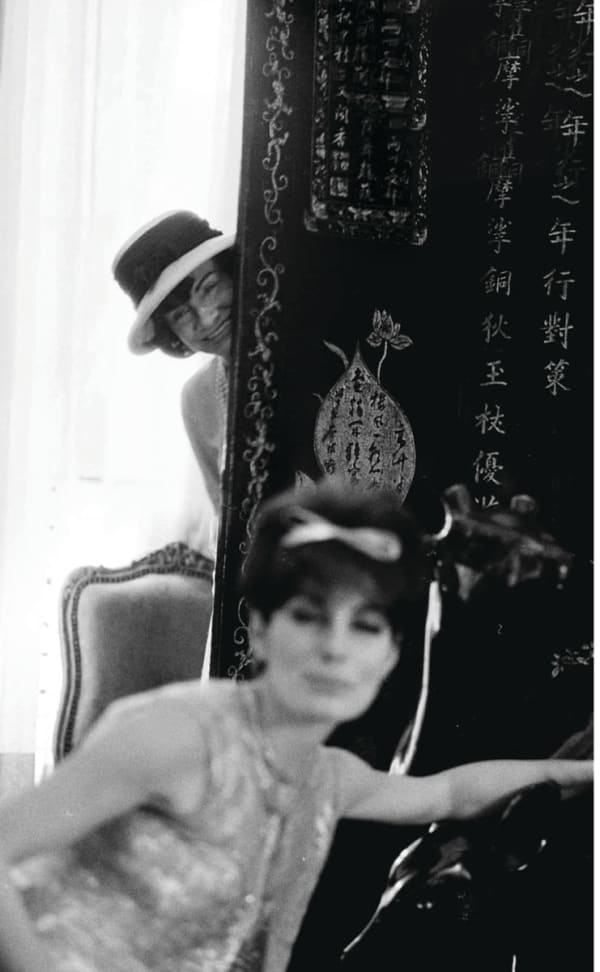 Black and white photograph of Designer Coco Chanel peeking from behind a screen with model Jackie Rogers in the foreground