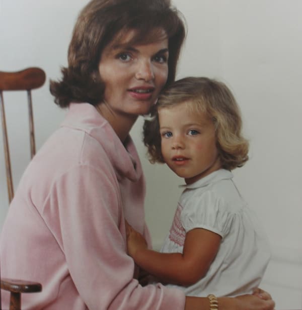 Color photograph of a young Caroline on the lap of her mother, Jackie Kennedy as they both turn their heads to face the camera