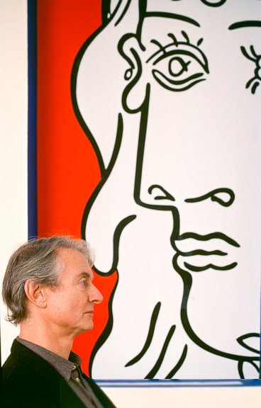 Photograph of Roy Lichtenstein in profile in front of a red, black, and white painting of a woman’s face