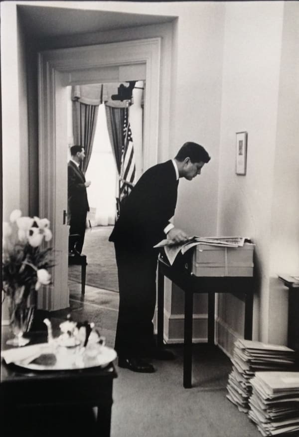 Black and white photograph of John F Kennedy standing and reading a newspaper in a stately interior during his visit to France