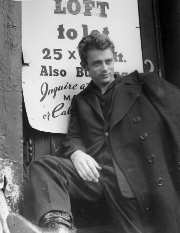 Black and white photograph of James Dean smiling in a black pea coat and propping one leg up on a stoop railing in front of a poster reading, "Loft to let"