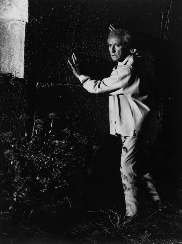 Black and white photograph of Jean Cocteau standing in grass with his hands against a wall of a building
