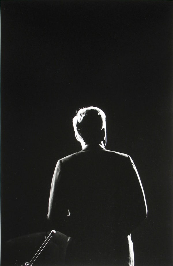 Black and white photograph of John F Kennedy's silhouette from behind, made visible against the black background by a thin halo of light outlining his figure
