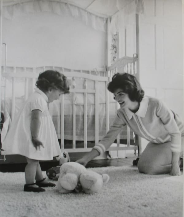 Black and white photograph of Jackie Kennedy in a nursery room motioning to a teddy bear on the floor in front of a young Caroline Kennedy, who looks down at the bear and is wearing a white dress