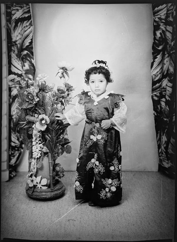 1960s black & white photograph of a young girl wearing an embroidered dress, in a photography studio with artificial flowers