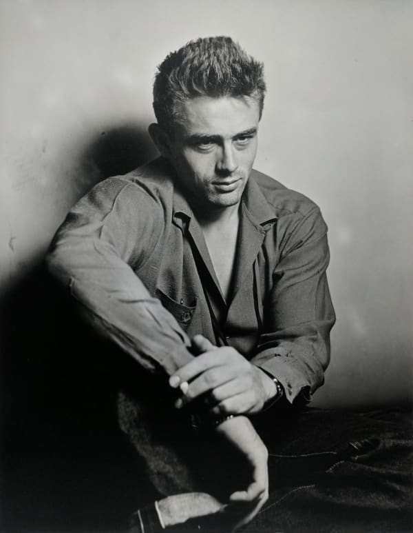 Black and white photograph of James Dean sitting against a wall with the crook of one arm resting on his knee and a cigarette between his fingers