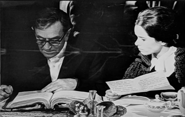Black and white photograph of Geraldine Chaplin as Tonya wearing a large black coat with soviet propaganda posters behind her, on the set of “Doctor Zhivago”