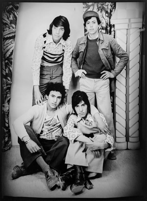 1960s black and white photograph of four young men wearing western attire, two men sitting on the floor with their arms wrapped around each other and two men standing