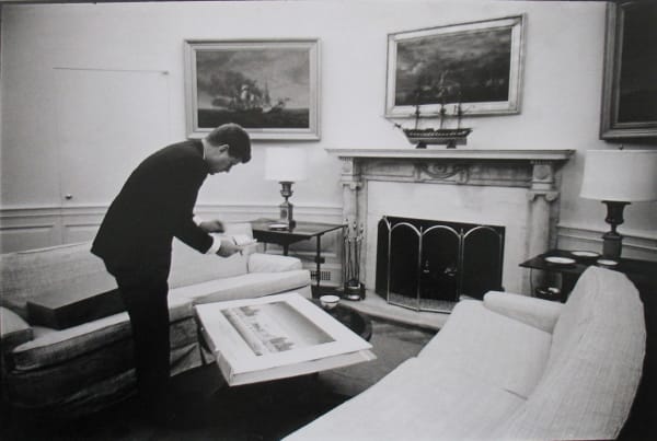 Black and white photograph of John F Kennedy bending over a painting in a room in the White House featuring paintings, furniture, and a fireplace