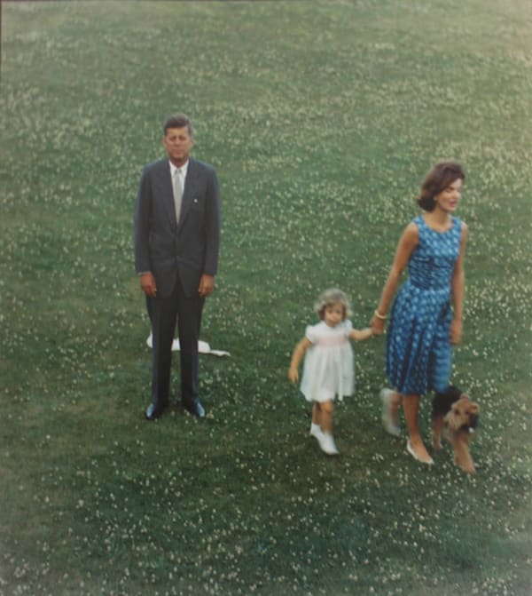 Color photograph from a high vantage point of John F Kennedy standing on a lawn while Jackie, Caroline, and a dog walk away from him