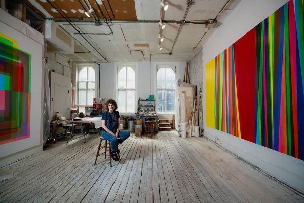 An artist, Betsy Kaufman, sits in an airy, sunlit loft setting surrounded by colorful abstract paintings
