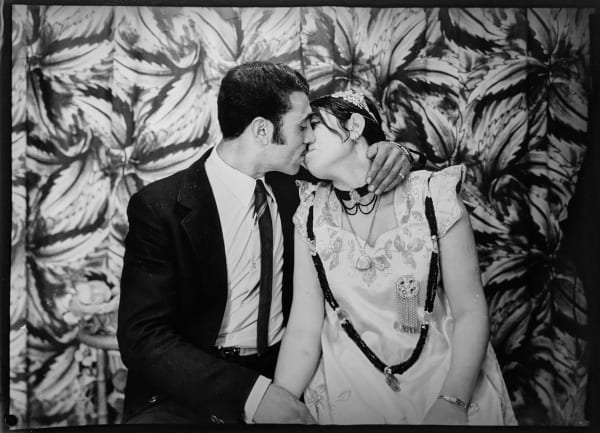 1960s black and white photograph of a young couple sitting and kissing while holding hands. The man is in a suit and the woman is wearing a white dress