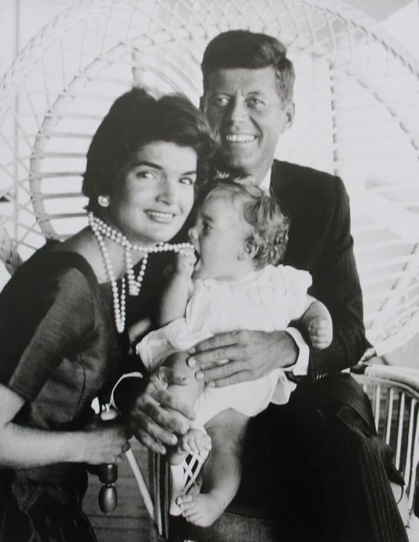 Black and white photograph of Jackie and John F Kennedy holding baby Caroline Kennedy who is putting her mother's pearls in her mouth