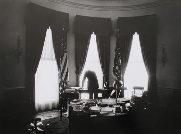Black and white photograph of John F Kennedy's silhouette bending over his desk in front of open windows in an otherwise dark Oval Office