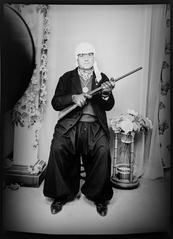 1960s black and white photograph of an older man sitting and holding a gun with a white head wrap on