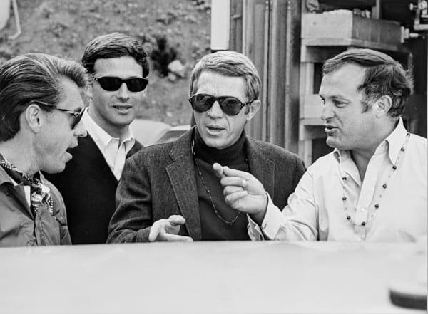 Black and white photograph of Steve McQueen, Peter Yates and crew sitting down outside speaking to each other, discussing the next scene, on the set of “Bullitt”