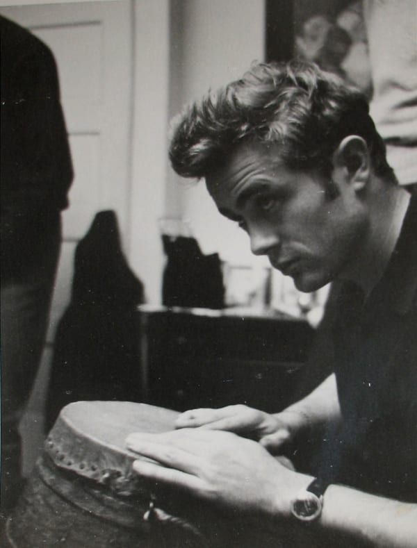 Black and white photograph of James Dean playing the bongos while glancing upwards in an apartment setting