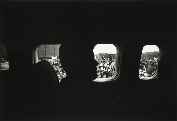 Black and white photograph of John F Kennedy's profile in silhouette in front of windows revealing a crowd of supporters outside his vehicle