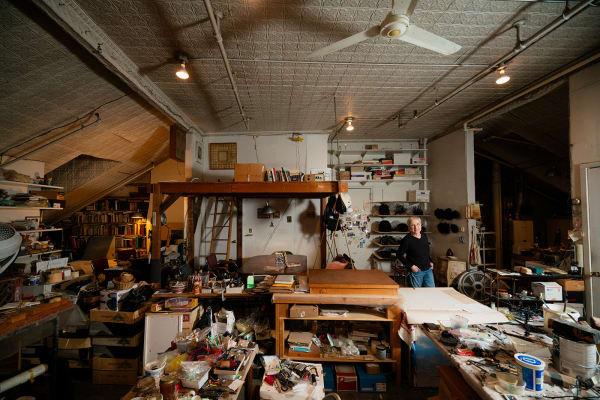 An artist, Gilda Pervin, standing amidst an array of work desks, art materials, and books in a loft setting