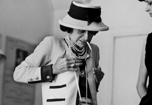 Black and white photograph of Designer Coco Chanel smoking a cigarette while looking down and patting herself