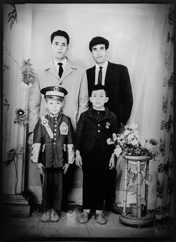 1960s black and white photograph of two adult men standing in suits with two young boys in front of them, one wearing a police uniform