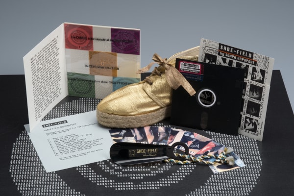 Sonya Rapoport Shoe-Field: Our Fate is on Our Feet, 1989 5.25 inch floppy disk; disk sleeve: photocopy and artist tape on paper; 5-page, accordion-folded booklet: photocopy, acetate collage, and artists’ tape on cardstock, 29.75 x 6.5 inches; printed gray cardstock insert 6 x 6.5 inches, 20 color photocopies mounted on cardstock, 1.625 x 2.5 inches each; acetate container, 6.75 x 7 inches; plastic shoe horn with gold print and yarn lanyard, 5.5 x 1.75 inches. With John O’Connell and John Watkins
