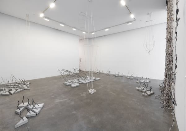 An installation view showing several arrangements of sculptures made of wire rope embedded into concrete bases as well as a curtain of twisted newspaper and a rope and wire sculpture hanging from the ceiling 