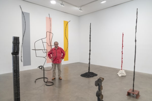 Installation View of Susan Inglett Gallery featuring Alain Kirili with several of his vertical sculptures and strips of gray, peach, and yellow on the wall behind him