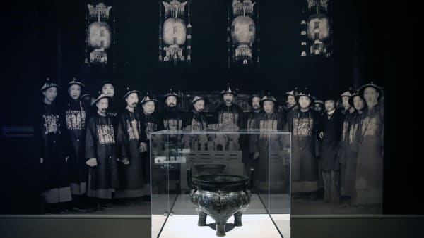 A bronze displayed in the exhibition hall, the background is a group of black and white photos of Qing Dynasty people on the wall