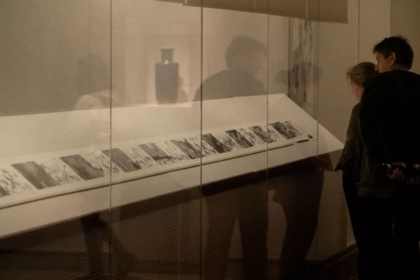 The album is unfolded and displayed in the museum's showcase, and there are two person look at it.