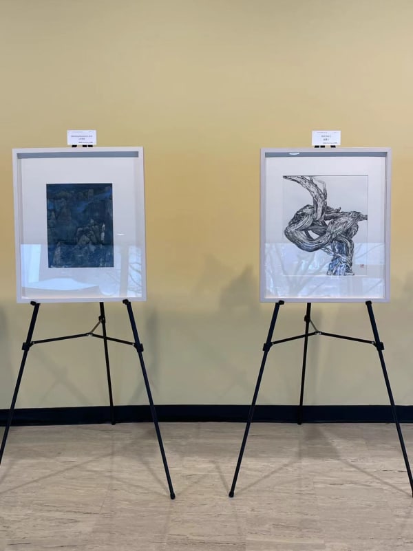 two artworks: one with black cirrus depicted on a piece of white paper, another depicting blue mountains and the sky exhibiting on a tripod stand